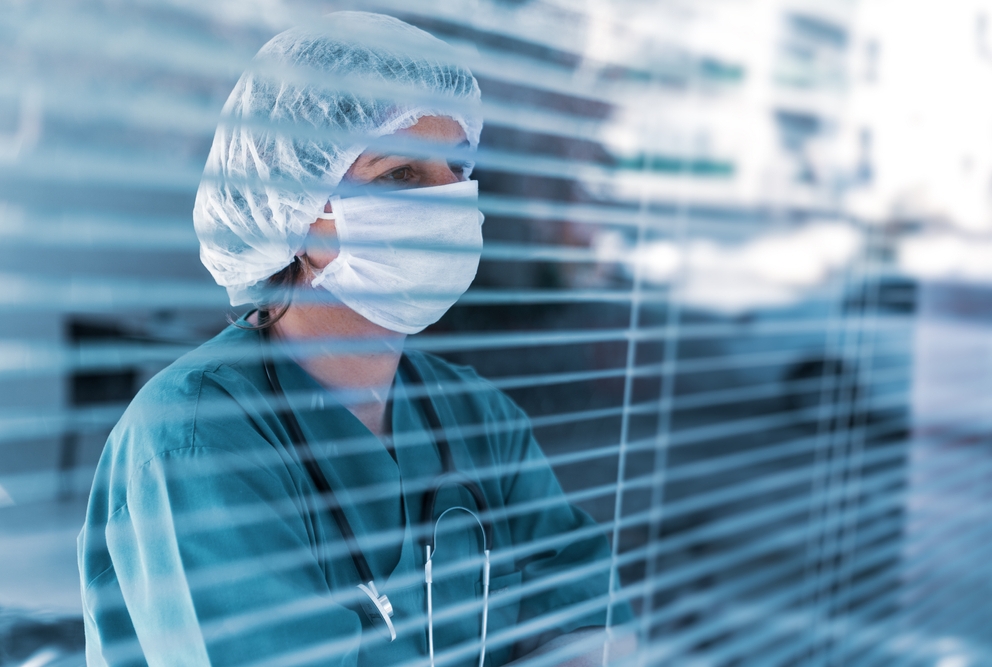 female doctor in scrubs and mask looking through window blinds
