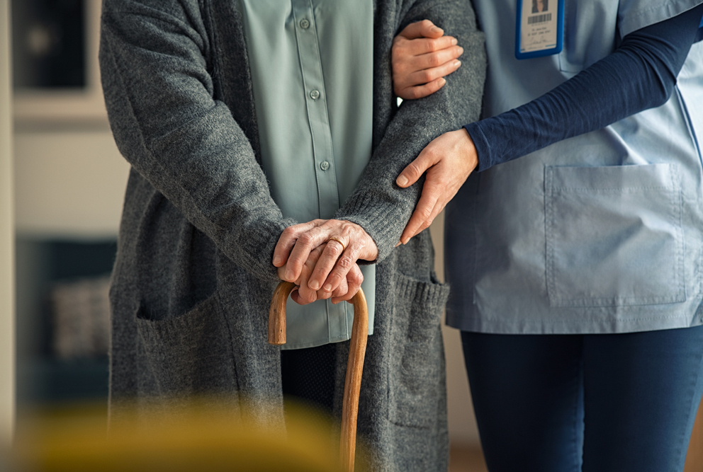 Elder woman using walking cane at nursing home with nurse holding hand for support.