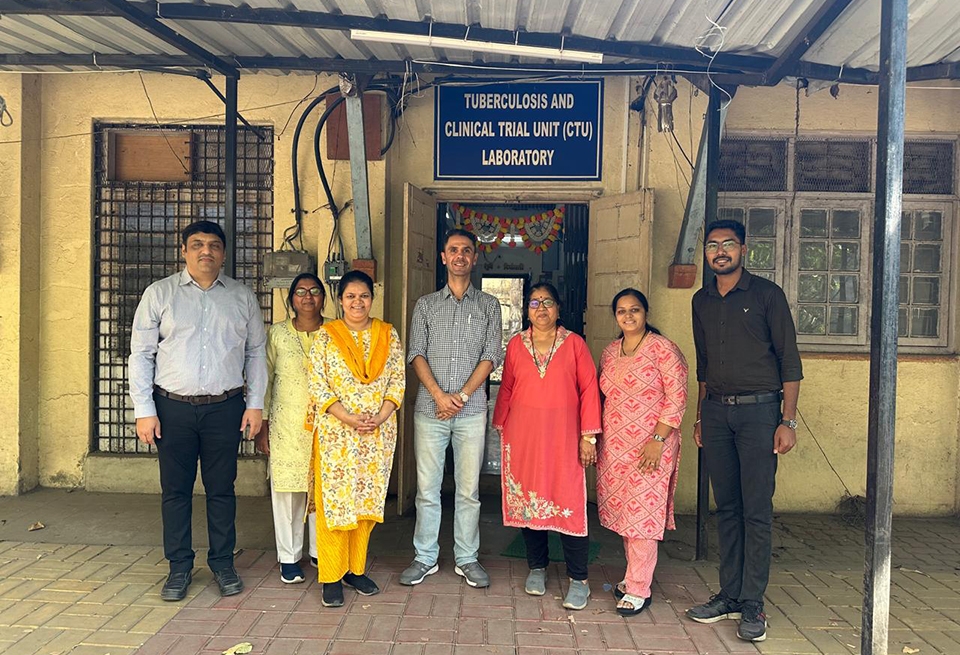 six people standing in front of a building with a sign reading &quot;Tuberculosis and Clinical Trial Unit (CTU) Laboratory&quot;