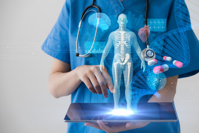 Person in blue scrubs holding tablet with digital images of skeleton, medications, DNA, among others displayed above its surface